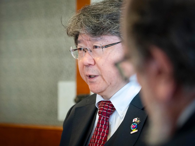A man in a suit and red patterned tie, wearing glasses, speaks indoors. Another person is visible in the blurred foreground.