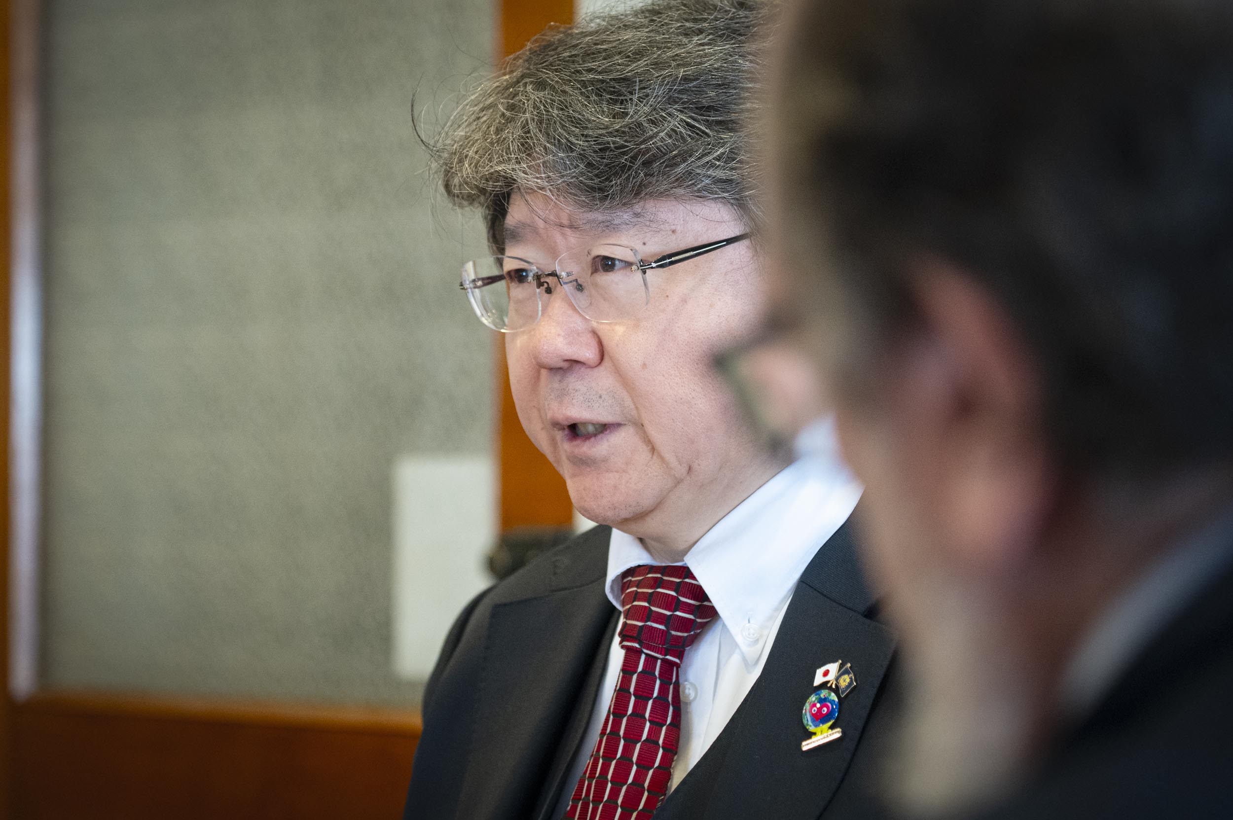 A man in a suit and red patterned tie, wearing glasses, speaks indoors. Another person is visible in the blurred foreground.