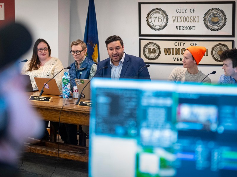 A panel of five people sits at a table during a meeting in Winooski, Vermont, with screens and audio equipment visible in the foreground.