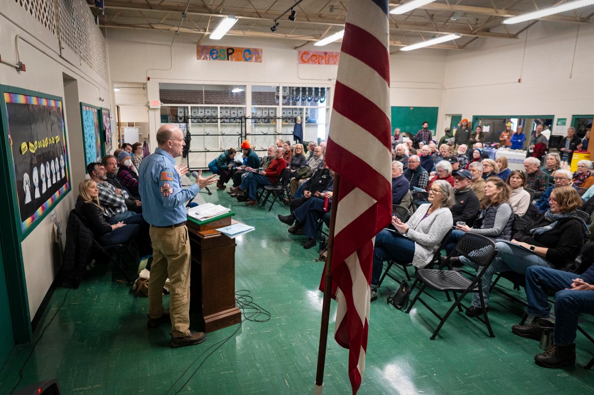 A man speaks at a podium to a seated audience in a school gymnasium, with an American flag in the foreground.