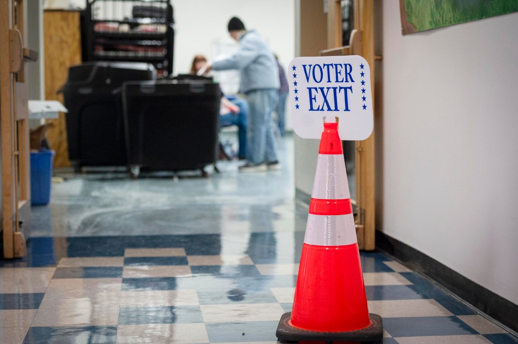 A traffic cone with a "Voter Exit" sign stands in a hallway; people are seated and talking in the background near voting booths.
