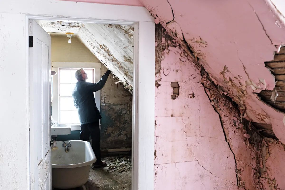 A person inspects a deteriorating attic bathroom with peeling paint, exposed plaster, and a freestanding bathtub.