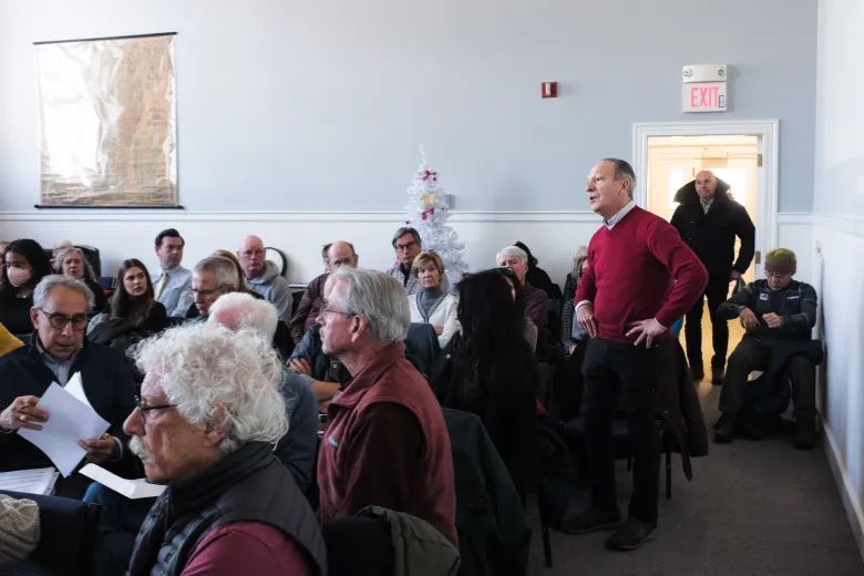 A group of people sit and stand in a meeting room; one man in a red sweater stands while others listen or look at papers.