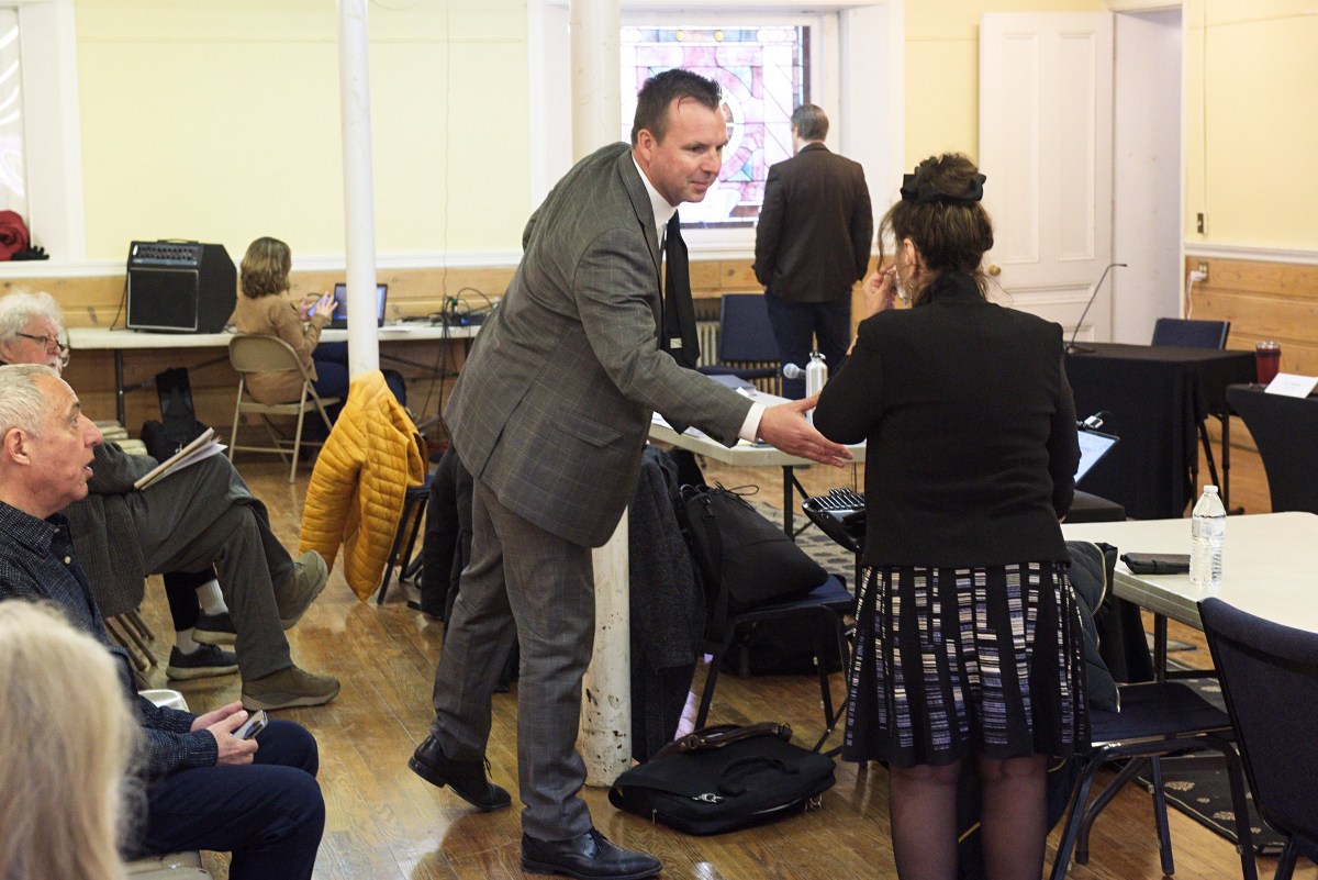 A man in a suit shakes hands with a woman in a black outfit in a meeting room, while other people are seated and working in the background.