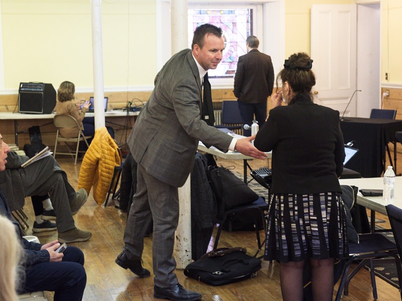 A man in a suit shakes hands with a woman in a black outfit in a meeting room, while other people are seated and working in the background.