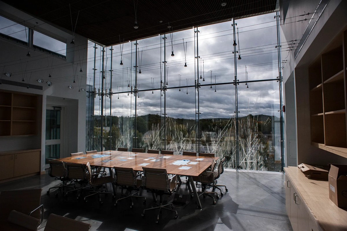 Modern conference room with a long table, office chairs, large glass windows, and an outside view of trees and cloudy sky.