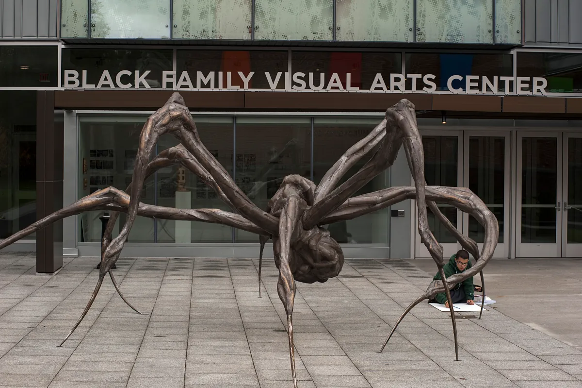 Large spider sculpture with long legs displayed outside the Black Family Visual Arts Center; a person is seated nearby on the ground, drawing or writing.