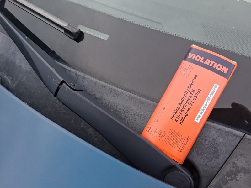A bright orange parking violation ticket is placed under the windshield wiper of a car on a wet windshield.