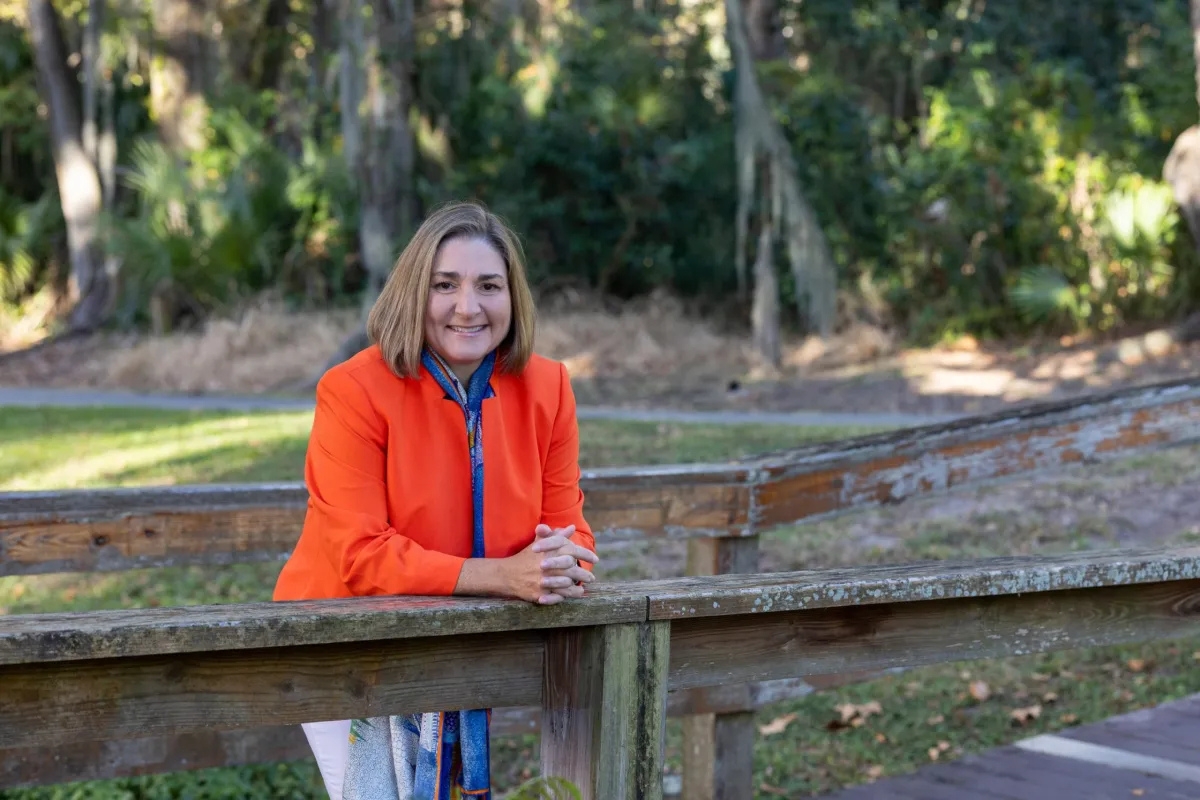A woman in an orange jacket stands outdoors, leaning on a wooden railing with trees and greenery in the background.