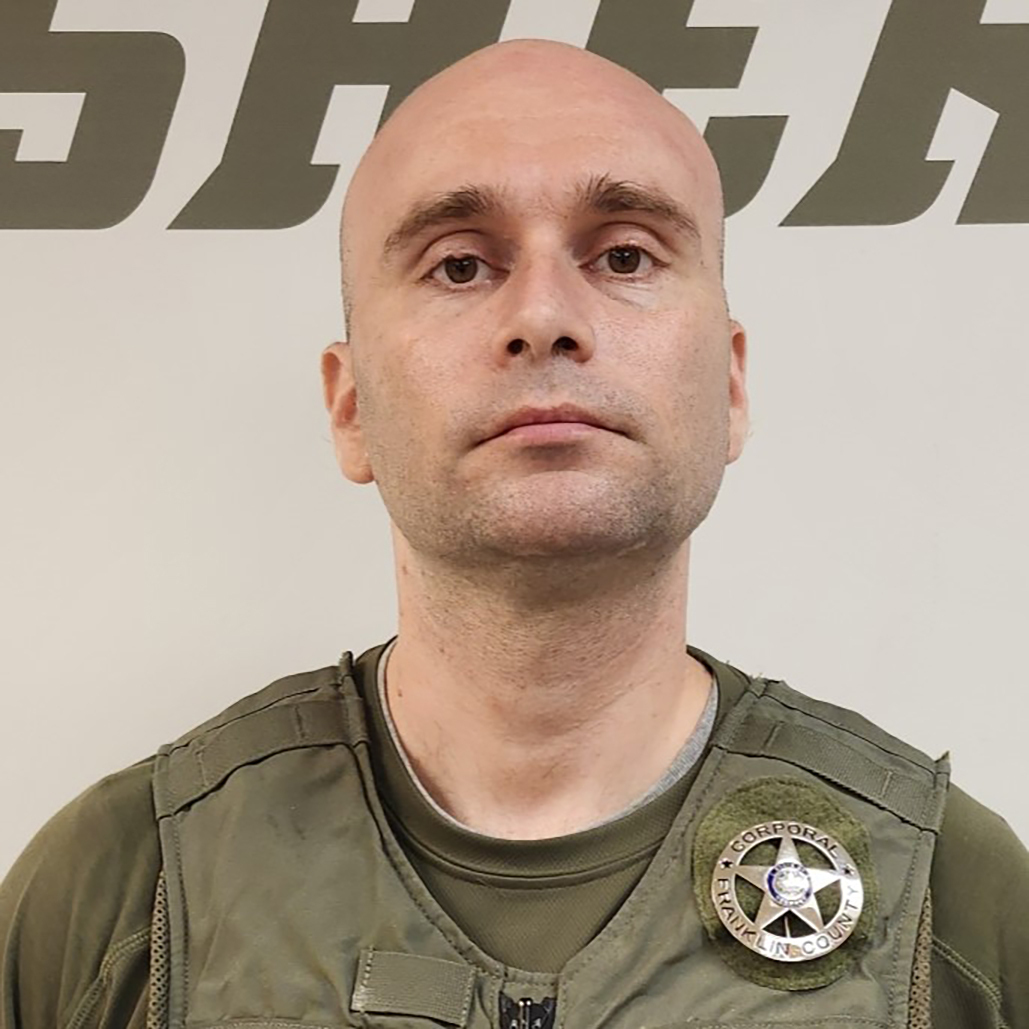 A man wearing a sheriff’s uniform and badge stands in front of a wall with large partial letters spelling “SHERIFF.”.