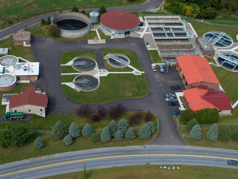 Aerial view of a wastewater treatment plant with circular and rectangular tanks, several buildings, driveways, and green landscaping.