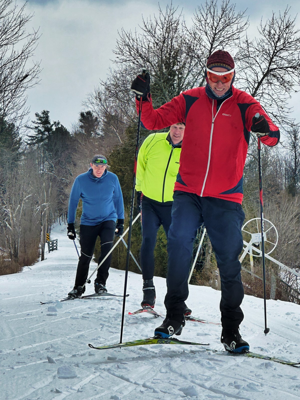 Three people cross-country skiing on a snowy trail surrounded by trees, dressed in winter gear, under a cloudy sky.