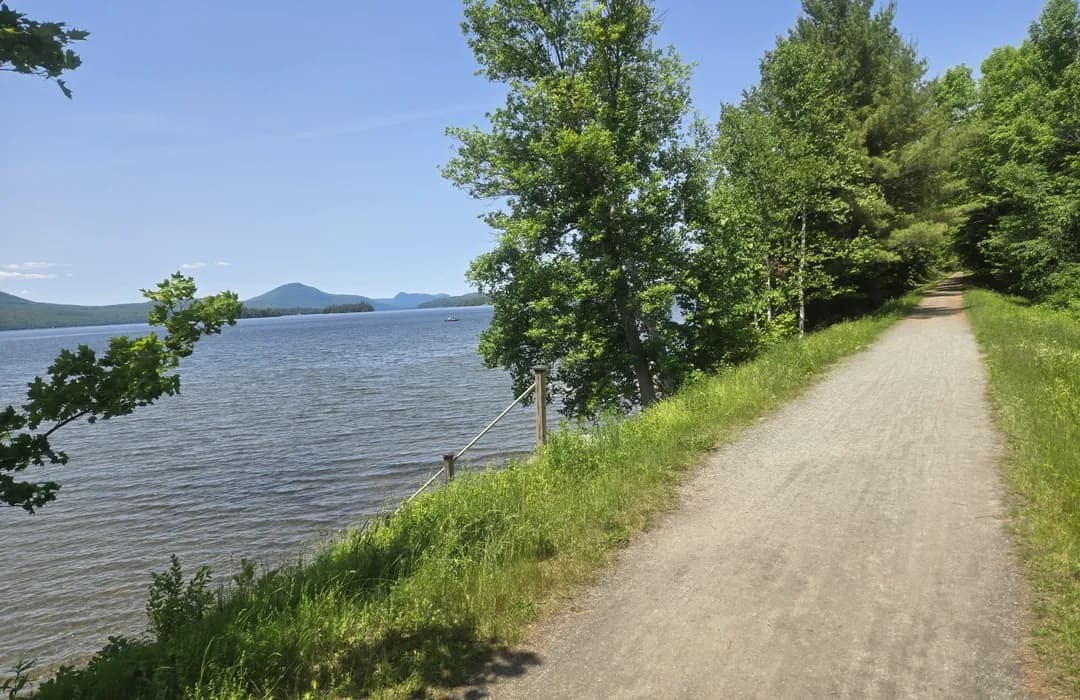 A gravel path runs alongside a lake bordered by green trees, with mountains visible in the distance under a clear blue sky.