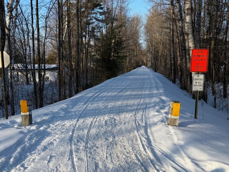 A snow-covered path through leafless trees is lined with yellow markers and signs indicating local snowmobile use only and speed limits.
