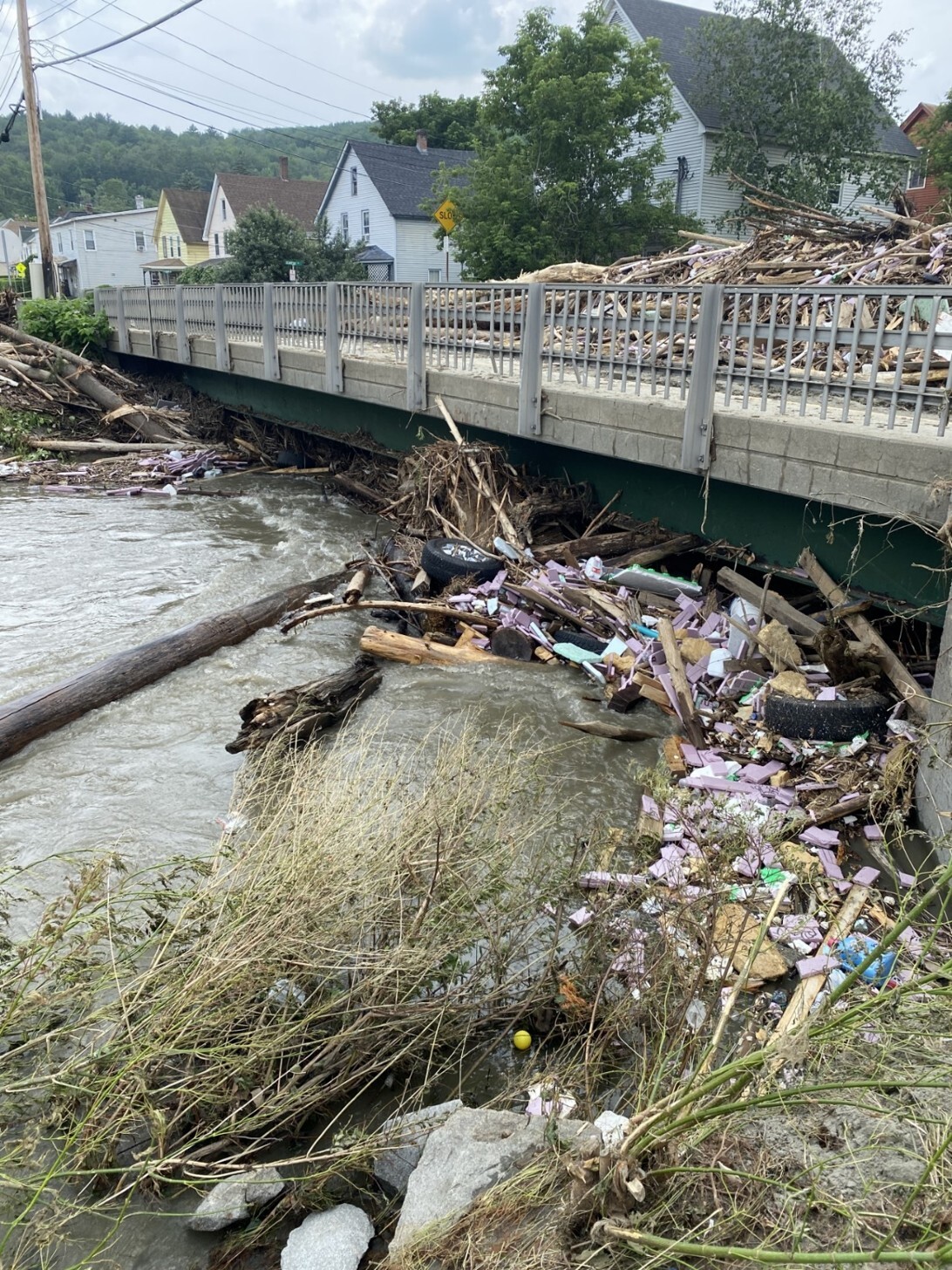 Flood debris, including wood, plastic, and vegetation, accumulates against a bridge over a swollen river; houses and trees visible in the background.
