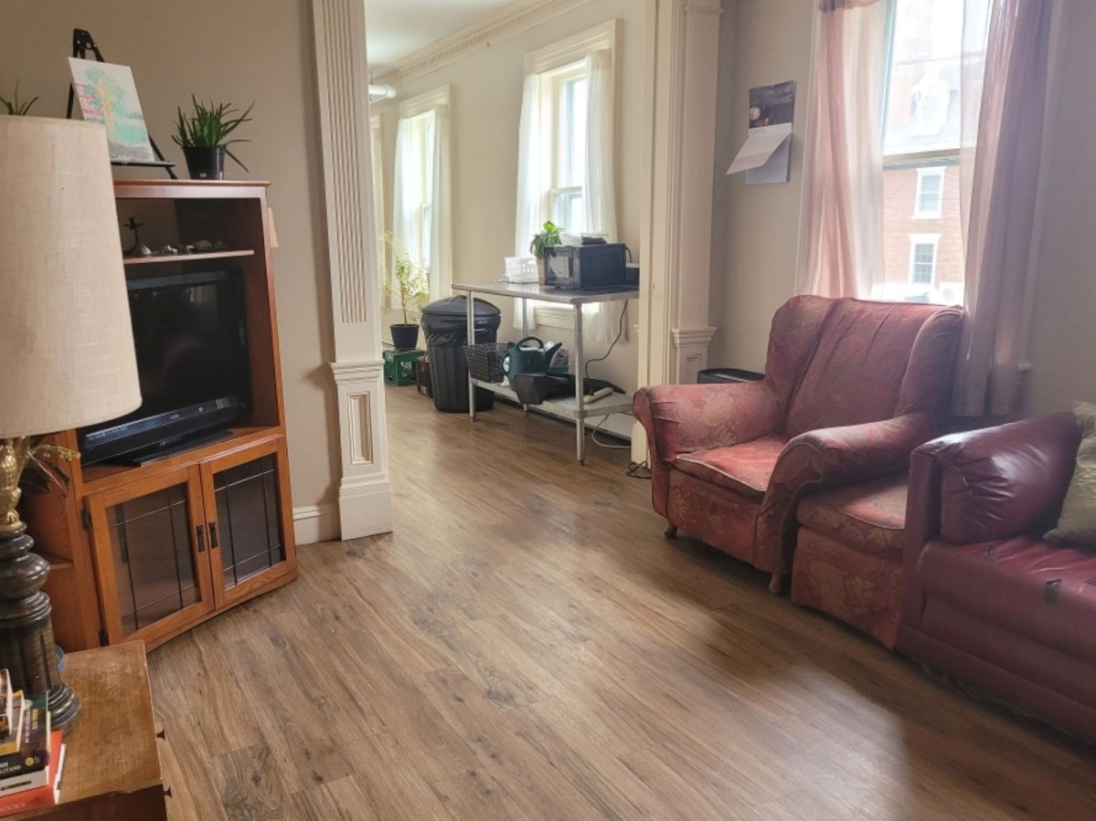 A living room with wooden floors, a red sofa and armchair, TV on a stand, and a table with office equipment near three windows.