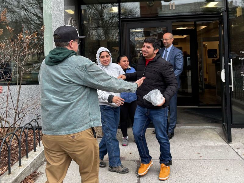Three people greet and smile outside a building entrance as a fourth person approaches them; a man in a suit stands in the doorway behind them.