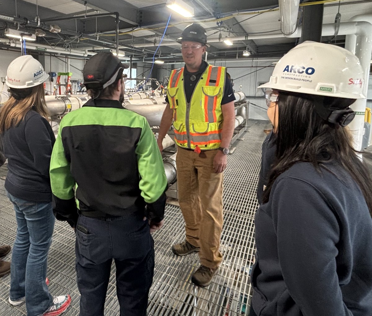 Four people wearing safety helmets and work attire stand in an industrial facility, having a discussion near metal pipes and equipment.