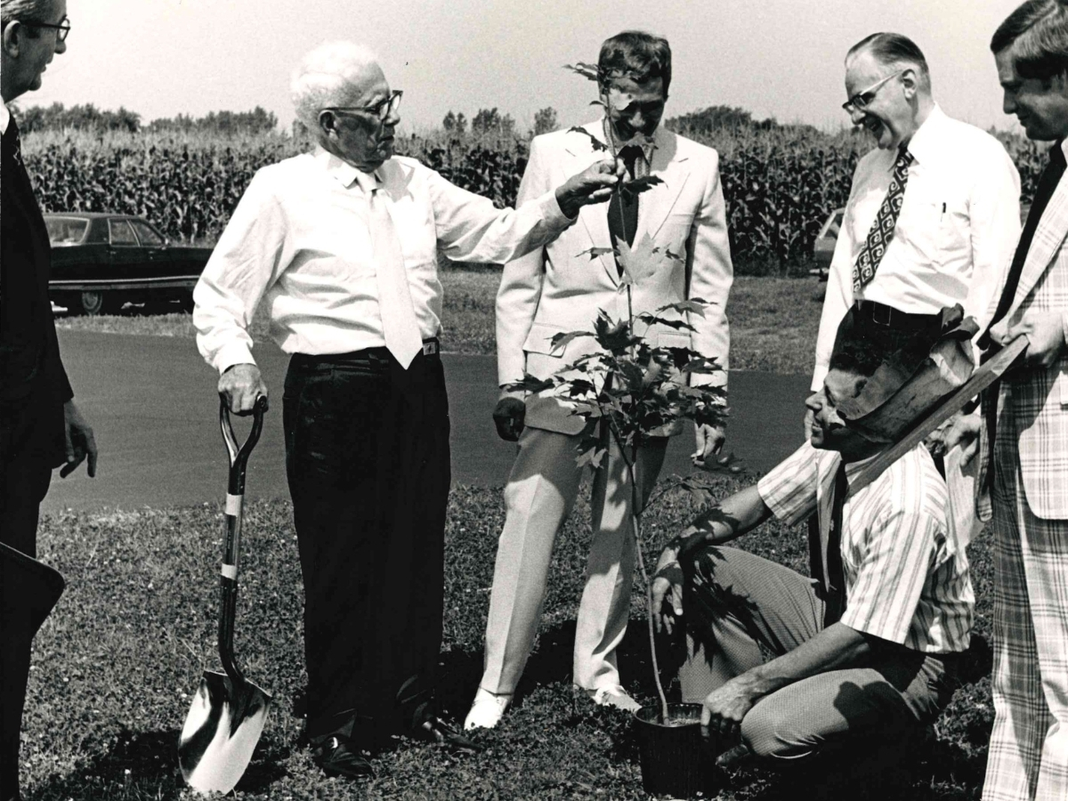Five men, four standing and one kneeling, participate in a tree planting ceremony outdoors. A shovel and small tree are visible in the foreground.