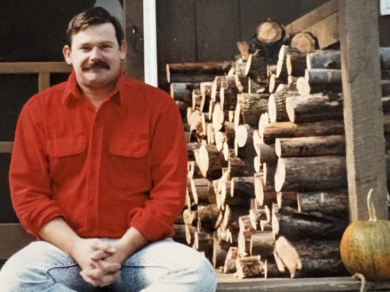 A man in a red shirt sits on a wooden porch next to a stack of firewood and a pumpkin.