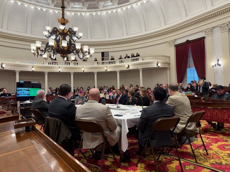 A large group of people attend a formal meeting in a domed room with a chandelier, red carpets, and balcony seating.