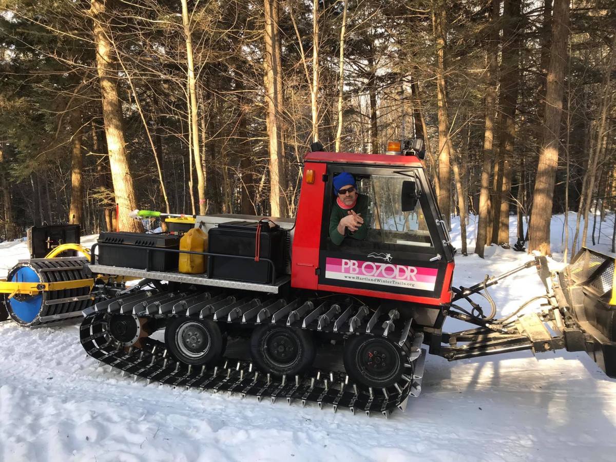 A person sits inside a red snow grooming vehicle parked on a snowy path in a forested area, surrounded by trees.