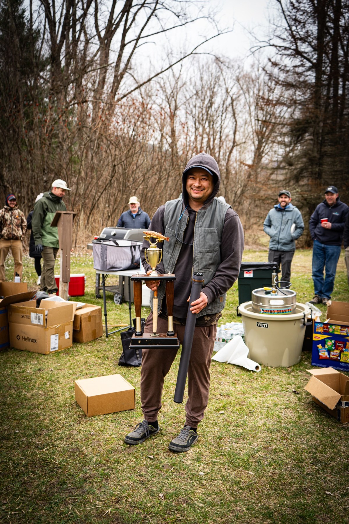A person in casual clothing holds a large trophy outdoors, surrounded by boxes and several people standing in the background on a grassy area with trees.