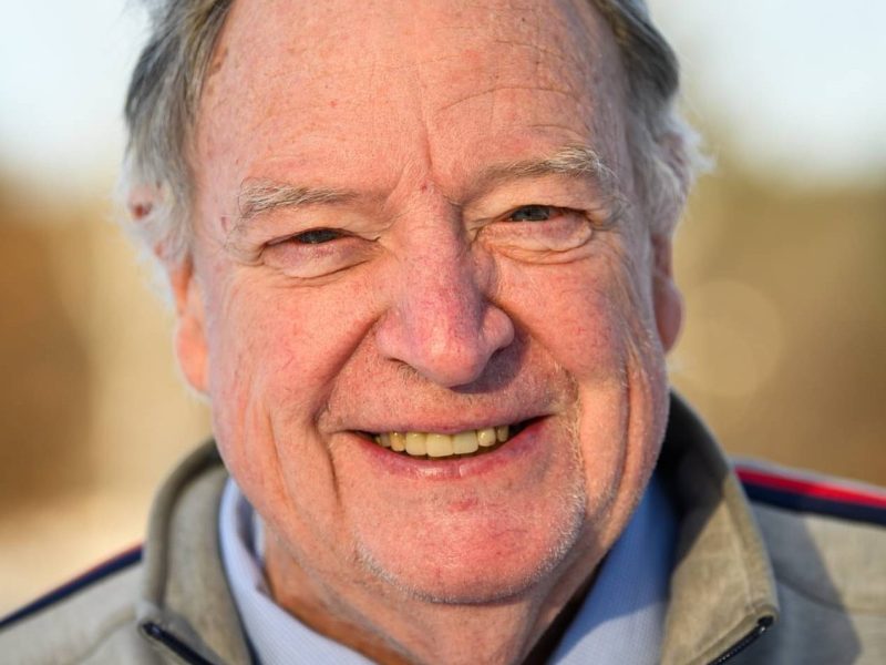 An older man with gray hair smiles at the camera outdoors, wearing a gray jacket over a blue shirt.