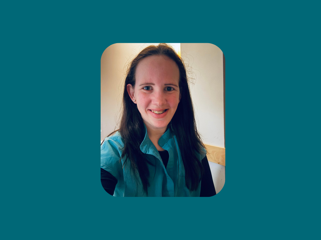 A person with long brown hair, wearing a teal shirt over a dark top, smiles at the camera indoors against a teal background with rounded corners.