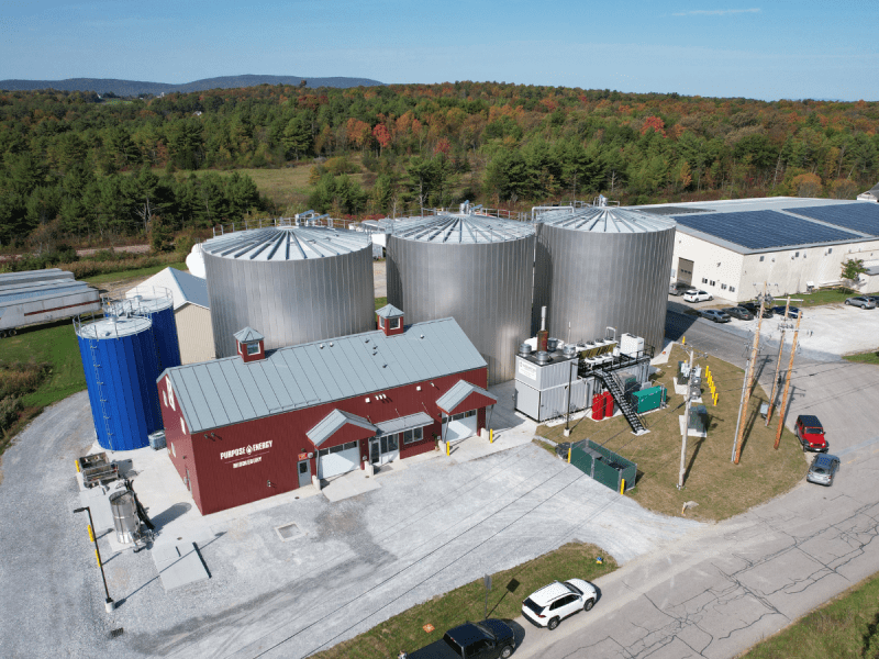 Aerial view of an industrial facility with large silver storage tanks, a red building, and surrounding trees under a clear sky.