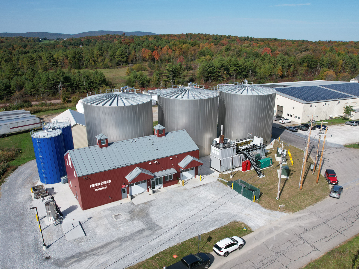 Aerial view of an industrial facility with large silver storage tanks, a red building, and surrounding trees under a clear sky.