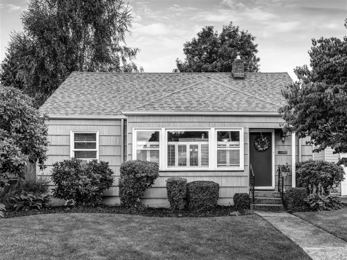 A single-story house with a shingled roof, front lawn, shrubs, and a red front door, photographed in black and white.