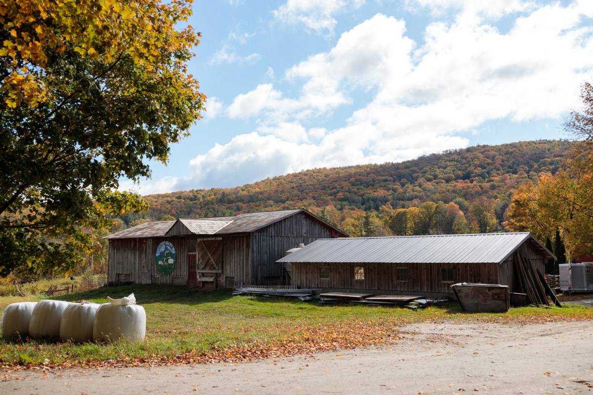 A rustic wooden barn with a metal roof sits in a grassy area, surrounded by autumn trees and hills under a partly cloudy sky. Round hay bales are visible in the foreground.