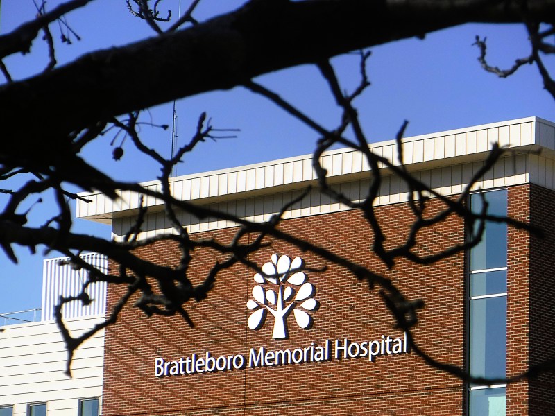 A building with a sign reading "Brattleboro Memorial Hospital" is partially obscured by tree branches against a clear blue sky.