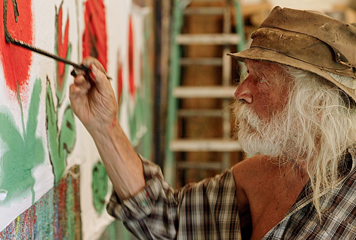 An elderly man with long white hair and a beard paints red flowers on a wall while wearing a plaid shirt and a weathered hat. A ladder is visible in the background.