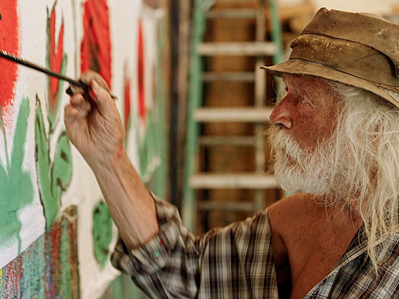 An elderly man with long white hair and a beard paints red flowers on a wall while wearing a plaid shirt and a weathered hat. A ladder is visible in the background.