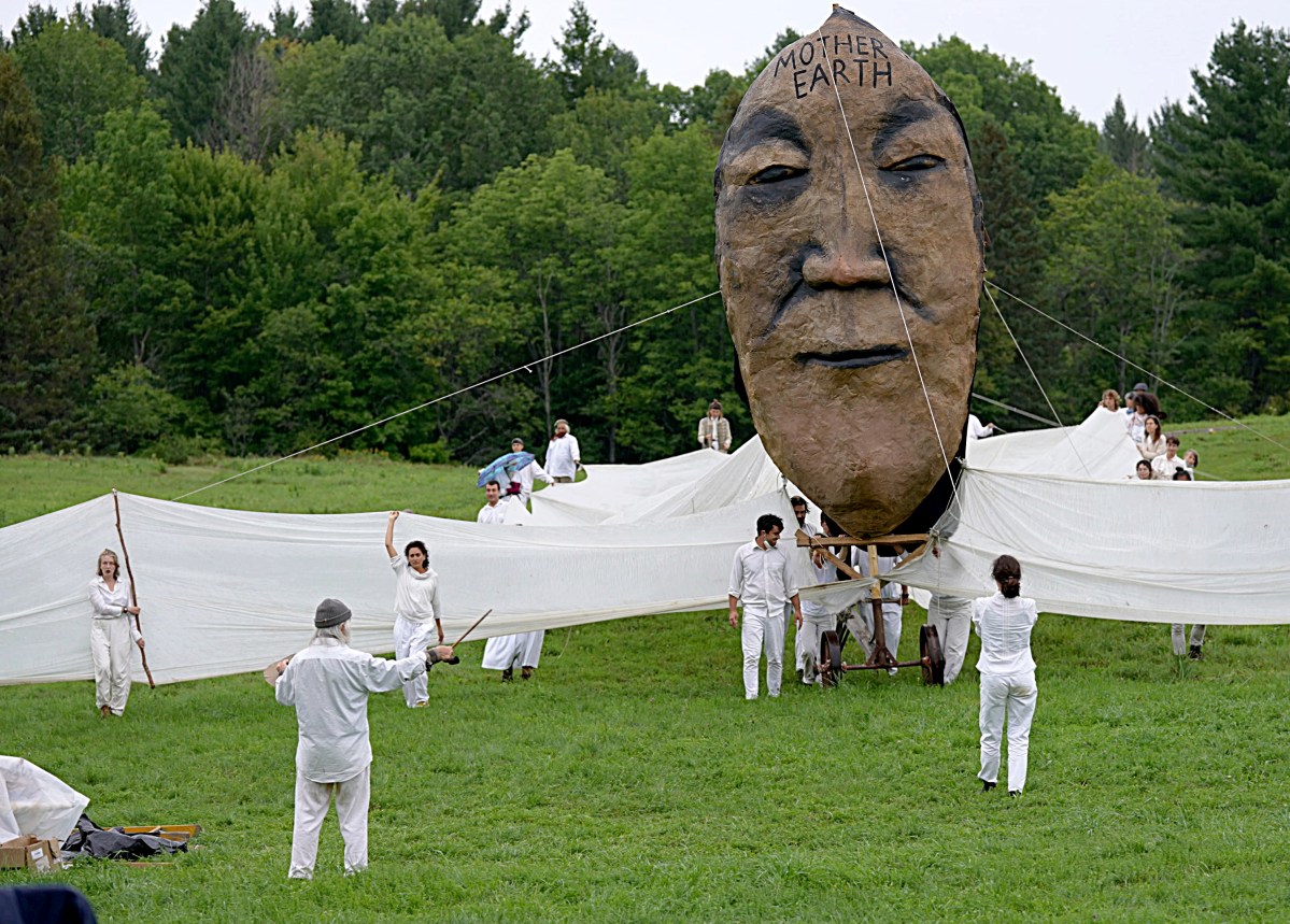 People in white clothing maneuver a large puppet with a human face labeled "Mother Earth" attached to long fabric wings in a grassy outdoor setting, surrounded by trees.