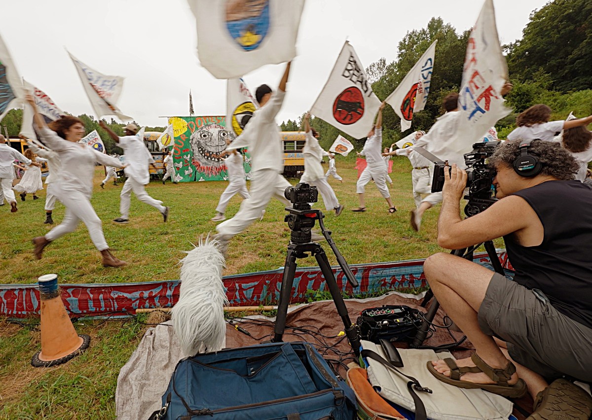 A group of people in white clothes wave flags while running past a cameraman filming the event outdoors, with colorful banners and trees in the background.