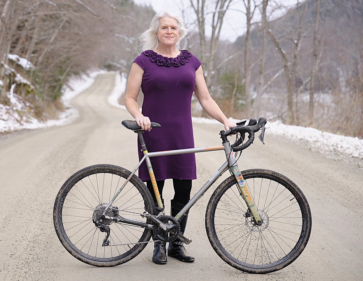 A person in a purple dress stands on a snowy dirt road, holding a bicycle by the handlebars. Trees line the road in the background.