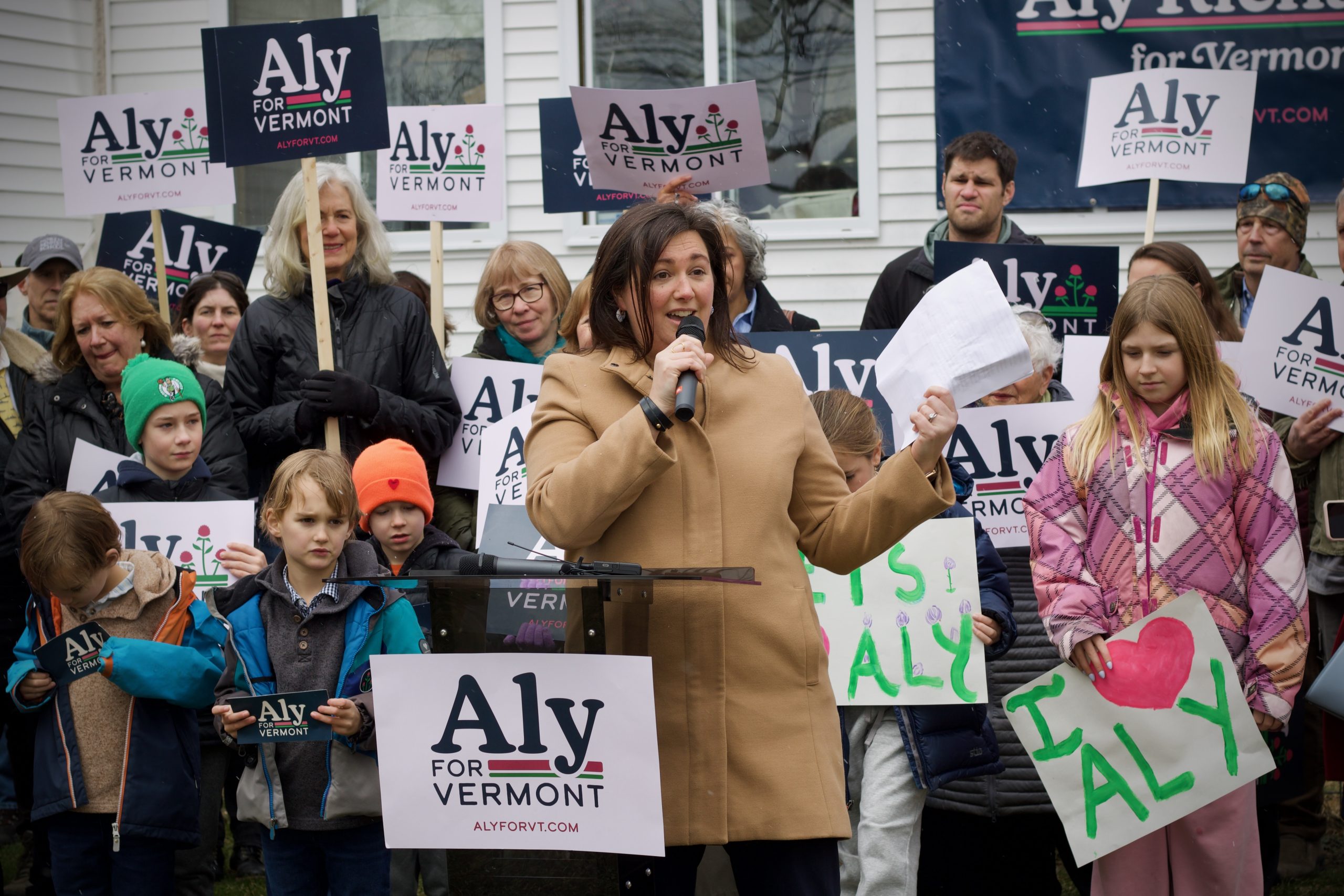 A woman speaks into a microphone at an outdoor rally, surrounded by people holding "Aly for Vermont" signs and handmade posters.