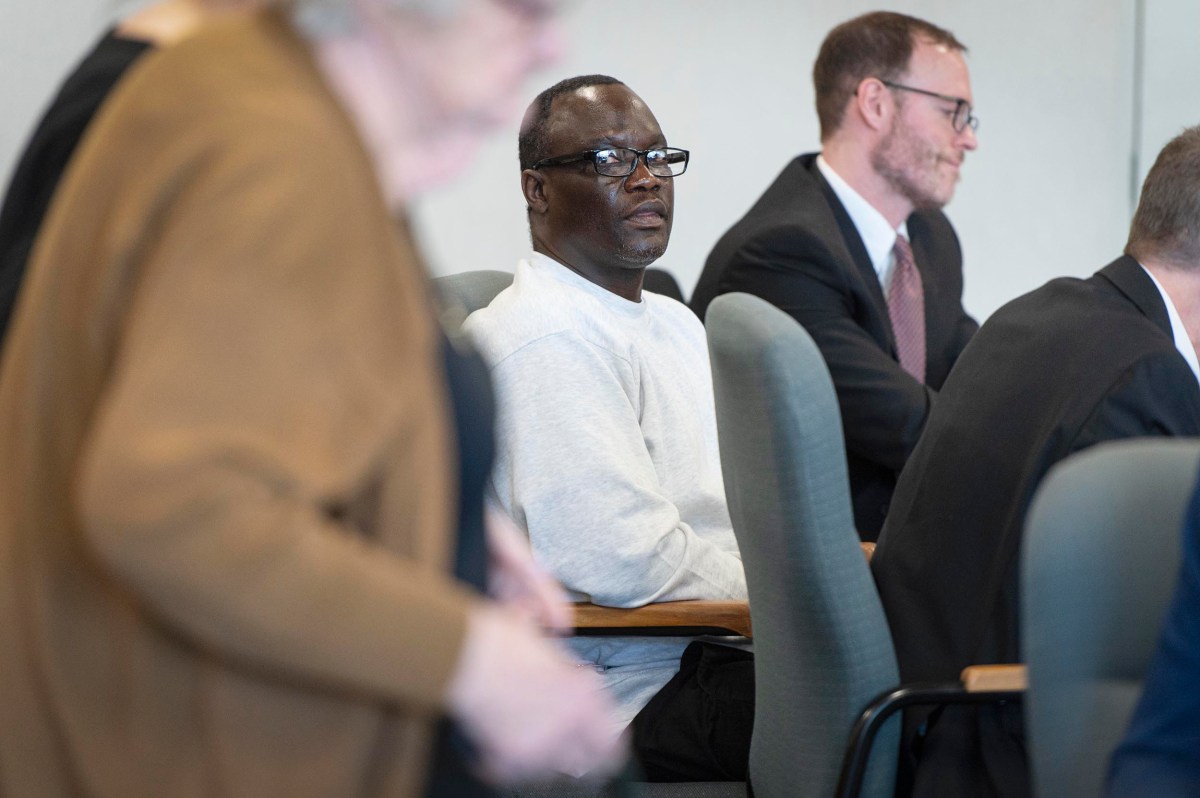 A man in a gray sweatshirt sits in a conference room with others, some standing and some seated, all appearing focused on a discussion or event.