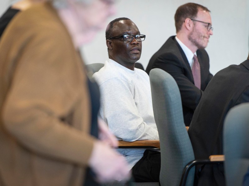 A man in a gray sweatshirt sits in a conference room with others, some standing and some seated, all appearing focused on a discussion or event.