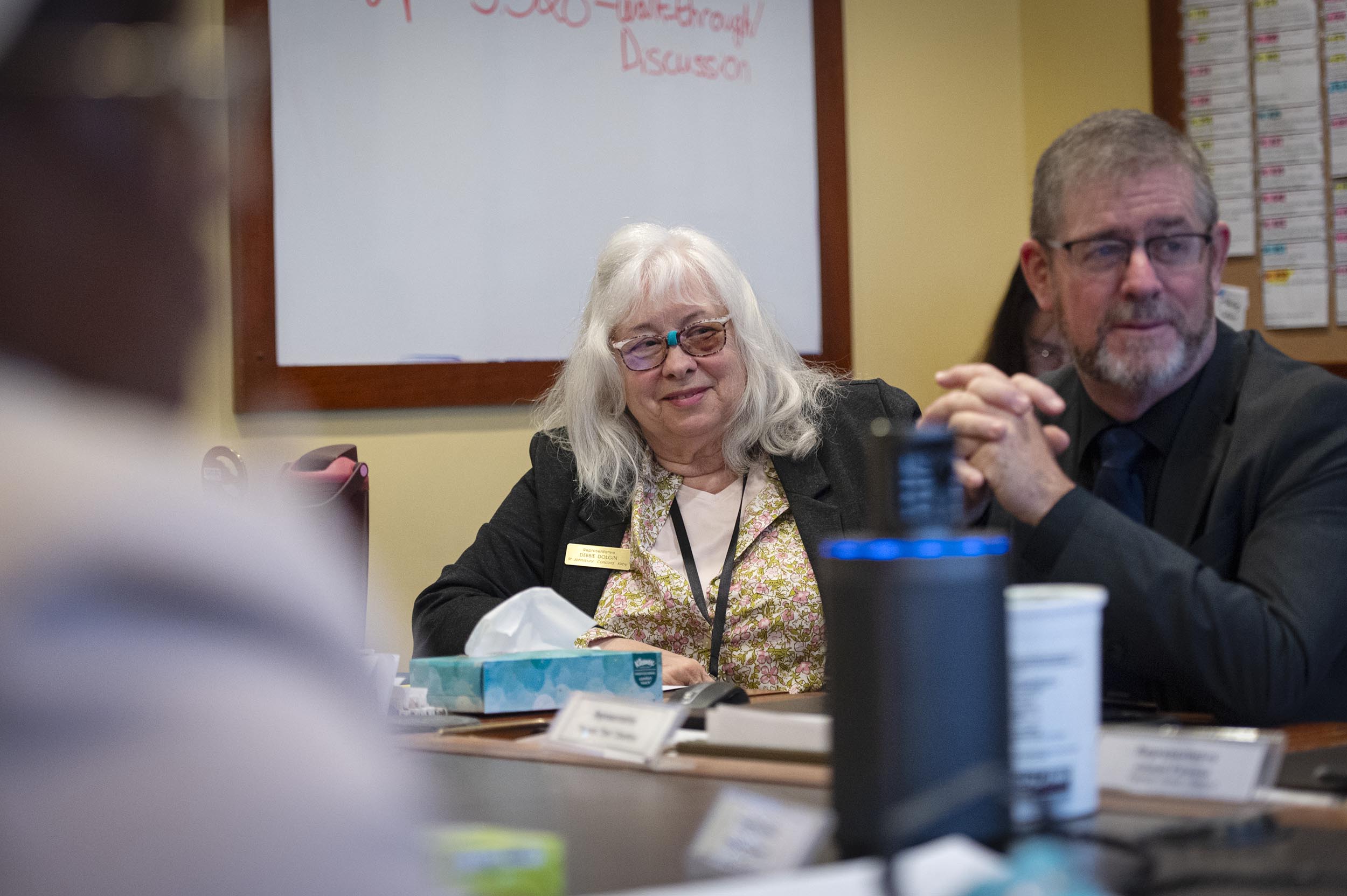Two people sit at a conference table during a meeting, with papers, a tissue box, and a cup on the table in front of them.