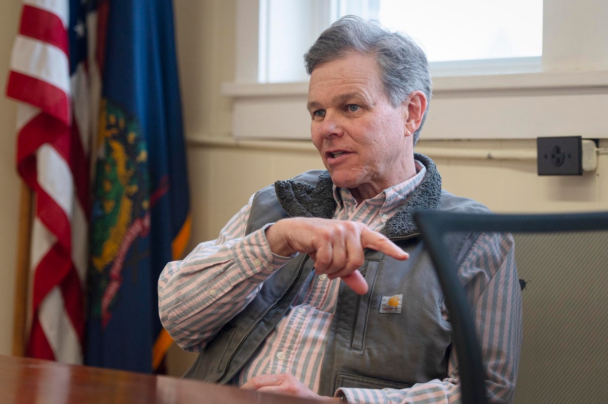 A man in a striped shirt and vest sits at a table, gesturing with his hand while talking. An American flag and another flag are visible in the background.