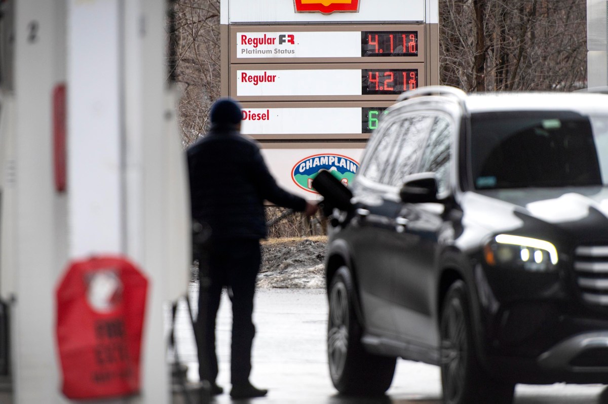 A person pumps gas into a black SUV at a gas station; fuel prices are visible on a sign in the background.