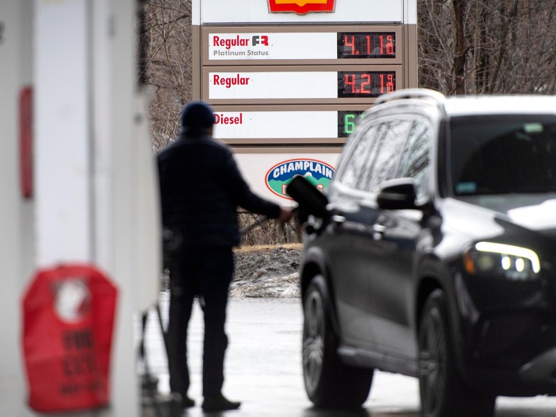 A person pumps gas into a black SUV at a gas station; fuel prices are visible on a sign in the background.
