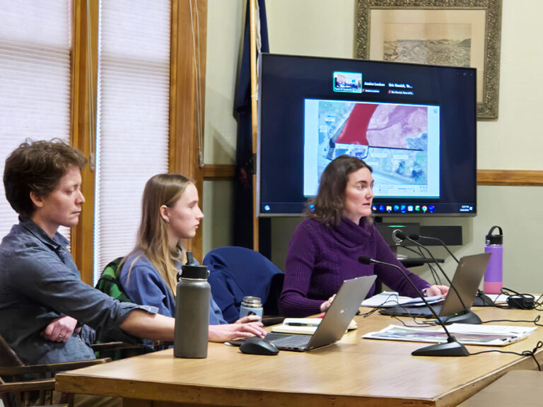 Three people sit at a table with laptops and microphones, while a woman speaks. A presentation with a map is displayed on a screen in the background.