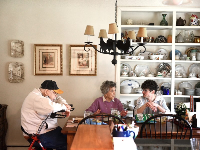 Three people sit around a dining table in a homey kitchen with shelves of dishes and decorative items in the background.