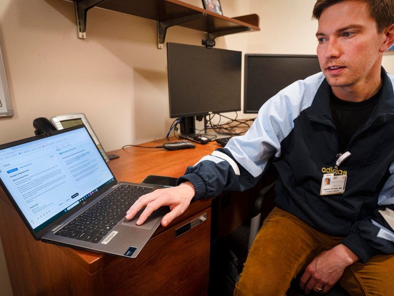 A person sits at a desk pointing to a laptop screen displaying a document, with two monitors and office items in the background.