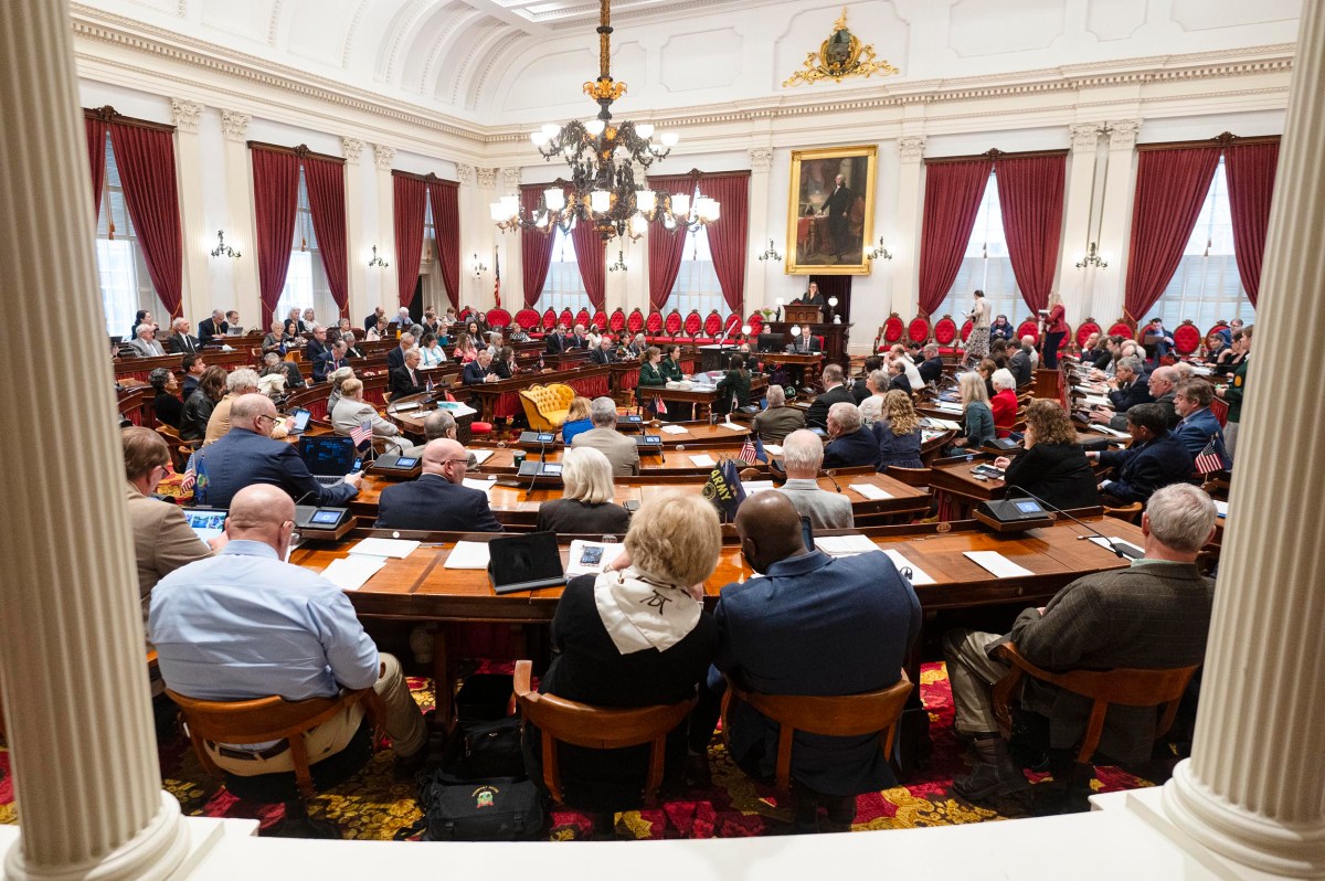 A large group of people sit at desks in a formal legislative chamber with red curtains, chandeliers, and a large portrait on the wall.